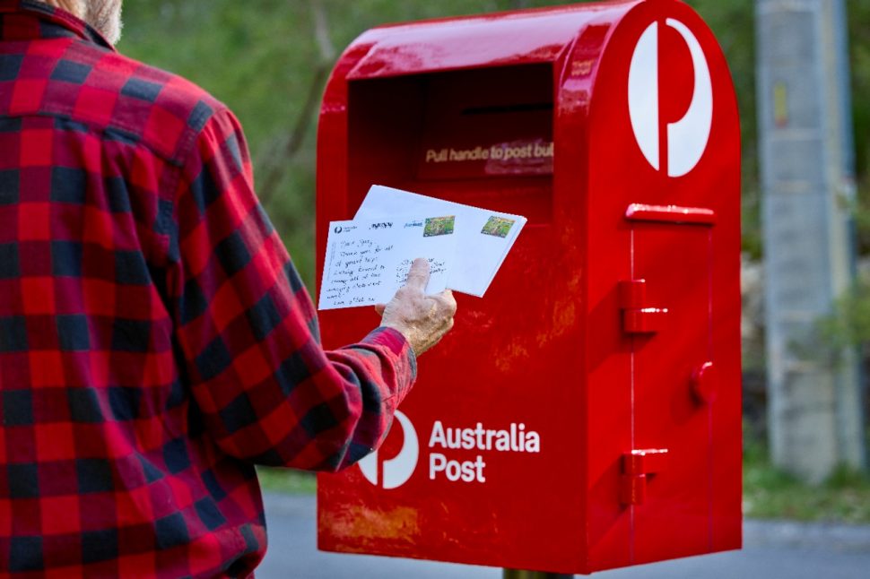 Person posting letter into a red Australia Post street mailbox.